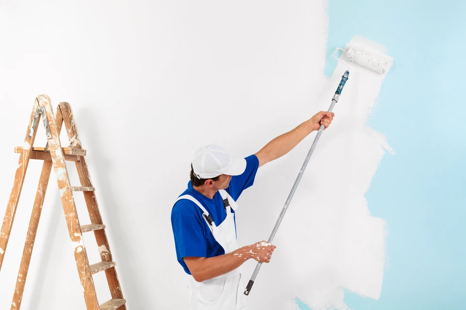 Peintre professionnel en combinaison bleue et casquette blanche applique de la peinture blanche au rouleau sur un mur intérieur