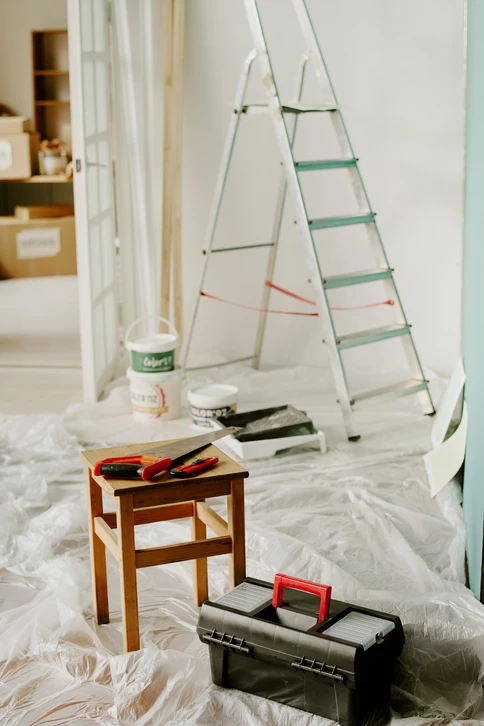 Peintre professionnel en combinaison blanche et casquette rouge tenant un pinceau, debout près d'une échelle pliante en aluminium contre un mur blanc intérieur en cours de peinture